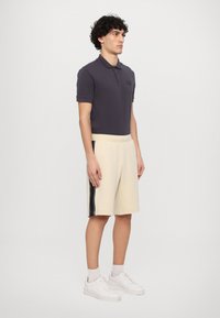 Camisa polo en gris oscuro con cuello y pequeño logo; pantalones cortos color crema con rayas negras a los lados; zapatillas blancas. Diseño simple y casual.