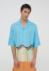 Camicia a maniche corte con bottoni di colore azzurro chiaro, caratterizzata da una stampa grafica di case colorate lungo il fondo. Tessuto leggero, vestibilità comoda.