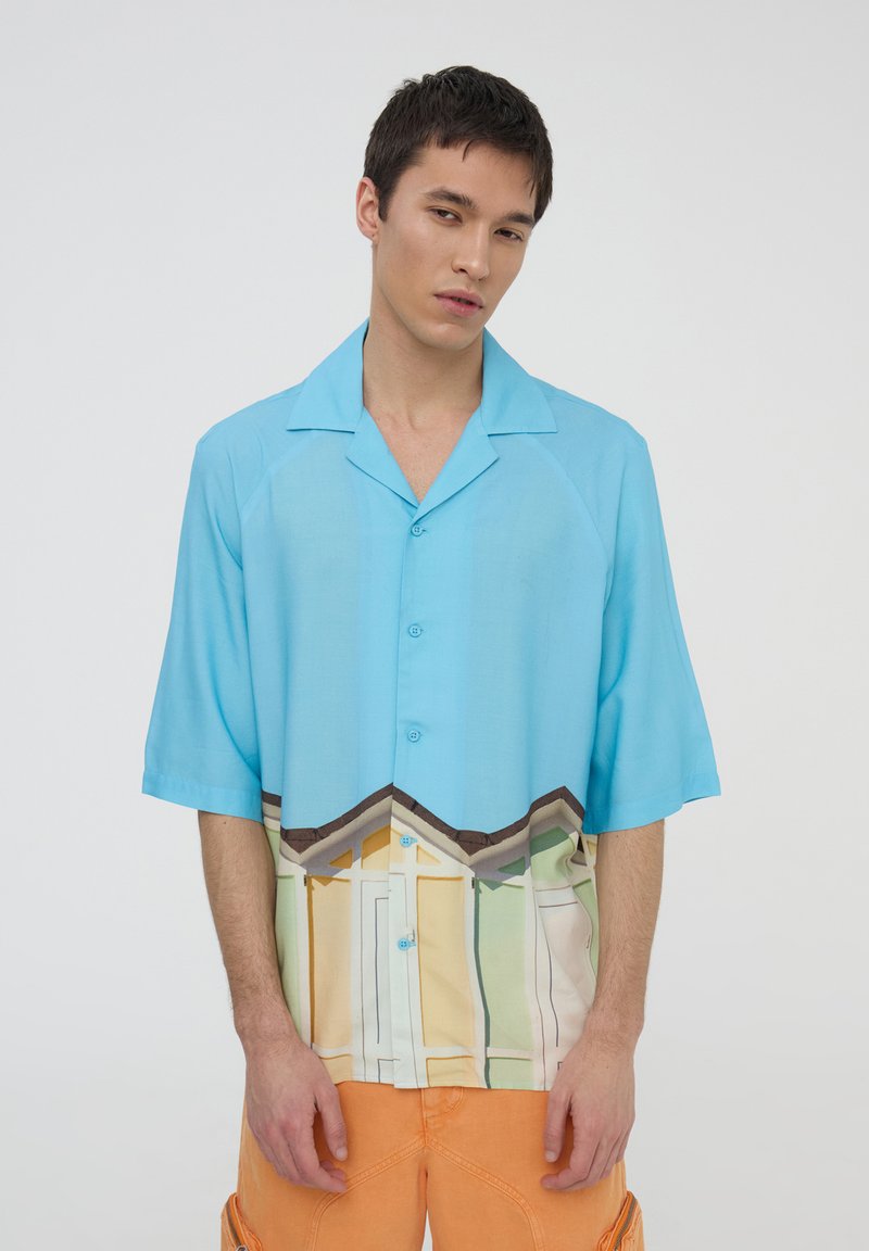 Camicia a maniche corte con bottoni di colore azzurro chiaro, caratterizzata da una stampa grafica di case colorate lungo il fondo. Tessuto leggero, vestibilità comoda.