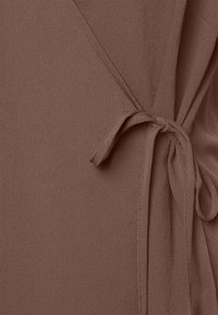 Tissu marron texturé avec une fermeture nouée, présentant un tombé doux et une finition lisse. Le design comprend des bords qui se chevauchent pour un style enveloppant.
