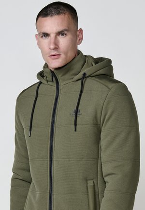 Sudadera con cremallera en color verde oliva, con tejido texturizado, capucha ajustable con cordón y detalles en negro. Cuenta con un bolsillo frontal y un logo en el pecho.