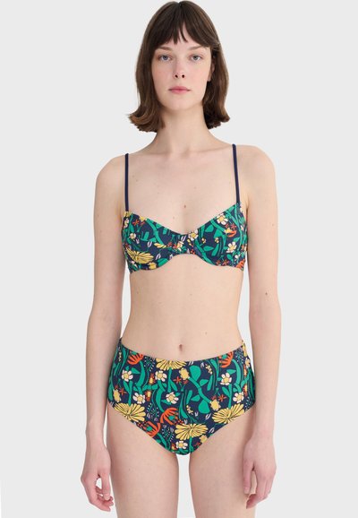 Femme portant un bikini bleu marine à motifs floraux jaunes, verts et orange, se tenant devant un fond gris clair uni.