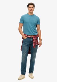 Korte mouwen teal T-shirt, blauwe jeans en beige sneakers, met een rood geruit overhemd om de taille geknoopt. Eenvoudige, casual outfit.