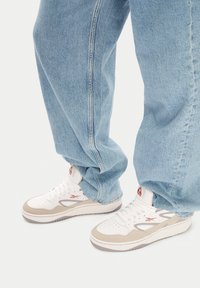 Biele a béžové tenisky s textúrovan�ým zvrškom, červenými logovými akcentami a pevným šedým podrážkou, skombinované s voľne strihanými svetlými modrými džínsami.