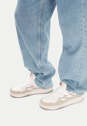Bílé a béžové tenisky s texturovaným svrškem, červenými logovými akcenty a pevným šedým podpatkem, doplněné volně střiženými světlými modrými džínami.