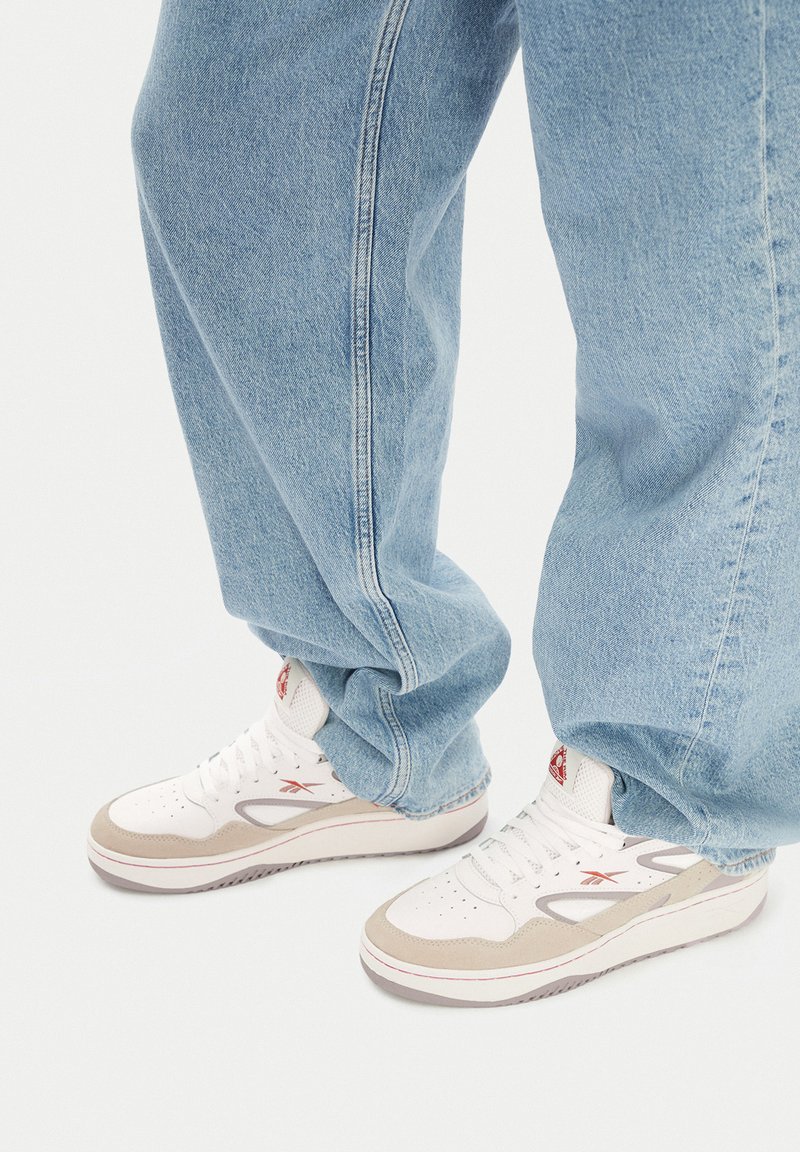 Biele a béžové tenisky s textúrovaným zvrškom, červenými logovými akcentami a pevným šedým podrážkou, skombinované s voľne strihanými svetlými modrými džínsami.