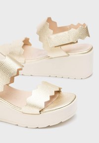 Sandalias de textura dorada con correas en zigzag, suelas de plataforma pronunciadas y acentos lisos. Presenta una base de color claro y una plantilla acolchada.