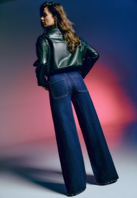 Veste en cuir noire au design court associée à un jean bleu foncé à taille haute et à jambes larges, avec poches arrière et ourlets effilochés.