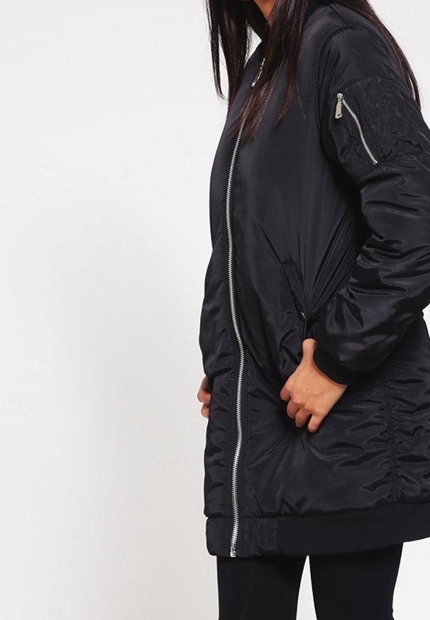 Femme portant une longue veste bomber noire avec fermeture éclair frontale, poches latérales et poche zippée sur la manche, debout devant un fond blanc uni.
