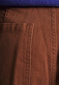 Bruna vävda byxor med en sidoficka, som har diagonala texturerade linjer. Överdelen visar en blå stickad tröja.