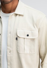 Beige overhemdd met een flexibele stof, voorzien van een klepzak met een zwarte knop en een klein logo-label aan de zijkant.