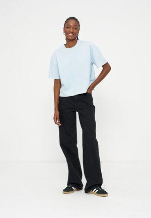 Jeune femme debout, main dans la poche, portant un t-shirt bleu clair, un jean noir à jambes larges et des baskets noires à rayures blanches.