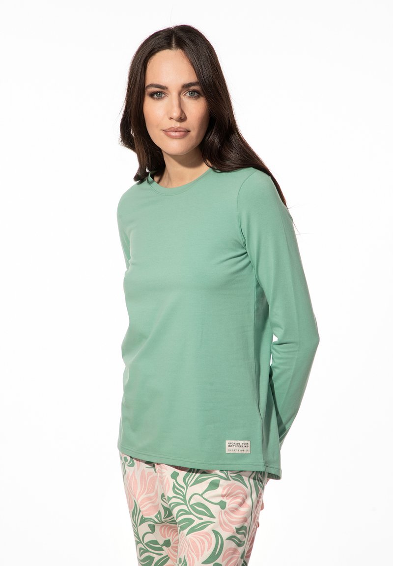 Helles grünes Langarmshirt aus glattem Stoff mit rundem Ausschnitt. Verfügt über ein kleines Etikett an der unteren Seite. Kombiniert mit floralen Hosen.