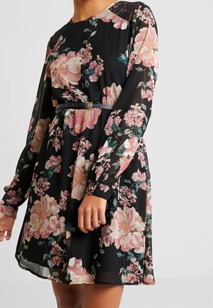 Frau trägt ein schwarzes Blumenkleid mit langen durchsichtigen Ärmeln und einem schmalen schwarzen Gürtel in der Taille, steht vor einem schlichten Hintergrund.