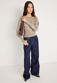 Μπεζ πλεκτό κάρντιγκ με κουμπιά και μακριά μανίκια, συνδυασμένο με φαρδιά τζιν παντελόνια σκούρου χρώματος. Κρατώντας μία καφέ δερμάτινη τσάντα. Σημειακά μαύρα παπούτσια.