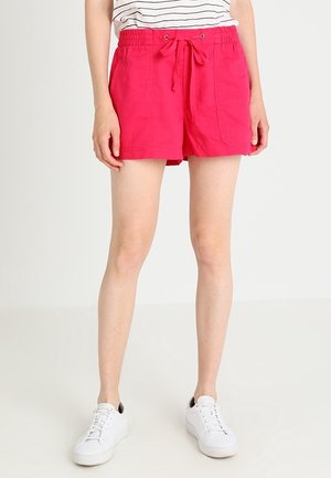 Kvinde iført lyse røde shorts med justerbar snor og forlommer, en hvid top med striber og hvide sneakers, stående mod en ensfarvet baggrund.