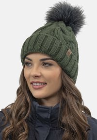 Green knitted beanie with a textured ribbed pattern and large faux fur pompom on top. Features a small brown logo tag on the side.
