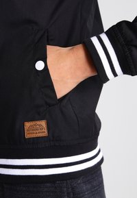 Chaqueta negra con puños de rayas blancas, que presenta una etiqueta de cuero marrón. El material es suave, con un ajuste ceñido y bolsillos laterales.