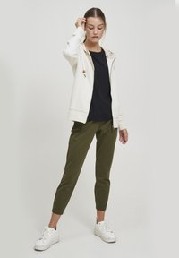 Lys beige zip-up hettegenser med dekorativ hette, svart t-skjorte og olivengrønn avsmalnende bukse. Inkluderer hvite sneakers med snøring.
