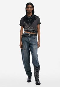T-shirt graphique noir avec des accents à franges, associé à un jean bleu ample. Des bottes de cowboy noires avec des détails de coutures décoratives complètent la tenue.