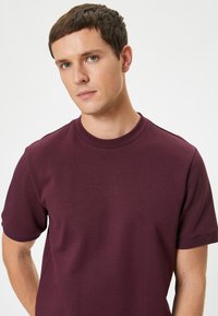 T-shirt a maniche corte con scollo rotondo di colore bordeaux scuro, realizzato in morbido cotone, con una silhouette aderente e polsini a coste.
