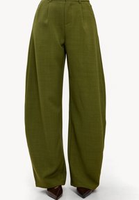 Pantalon à jambes larges en tissu vert olive, avec des plis sur le devant, une texture lisse et une ceinture cintrée. Chaussures à talons marron visibles.