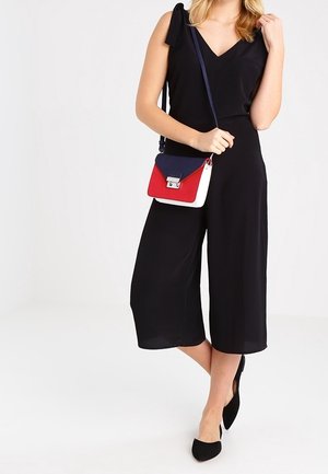 Sort ærmeløs jumpsuit med V-udskæring og skulderdetalje med sløjfe; kombineret med en tredelt crossbody-taske i rød, hvid og marineblå.