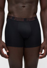 Buste masculin portant un boxer noir Puma avec une ceinture à logo, montré de la taille jusqu'au milieu de la cuisse sur un fond uni.