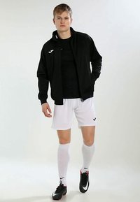Chaqueta atlética negra con cremallera, camiseta negra, pantalones cortos blancos, calcetas blancas hasta la rodilla y zapatos de fútbol negro y rojo con acentos del logo.