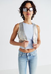 Zilverkleurige cropped vest over een lichtblauw geribbeld shirt en lichtblauwe jeans. Het vest heeft een knoopsluiting en geen mouwen.