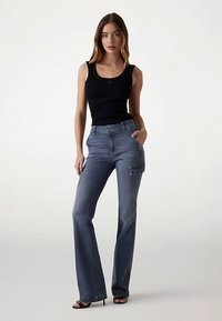 Jean en denim bleu foncé avec jambes évasées, doté d'une poche cargo latérale et de détails de couture subtils. Associé à un débardeur noir.