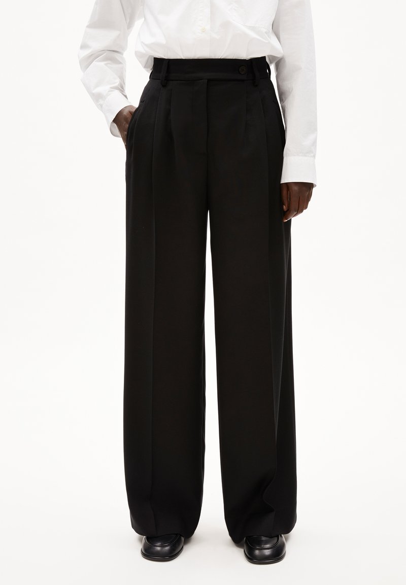 Zwarte wijde broek van gladde stof, met voorplooien en zijzakken. De taille heeft een knoopsluiting en riemlussen.