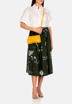 Camicia bianca a bottoni, gonna floreale verde scuro con motivi blu e bianchi, borsa a tracolla gialla e scarpe con tacco trasparenti.