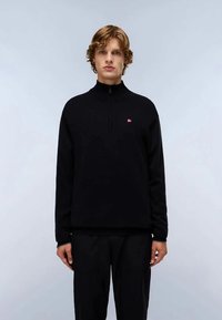 Un jeune homme avec des cheveux bouclés porte un pull noir à col zippé et un pantalon noir, se tenant devant un fond gris clair uni.