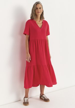 Kvinde med skulderlangt gråt hår iført en løs rød midi-kjole og brune sneakers, stående foran en hvid væg.