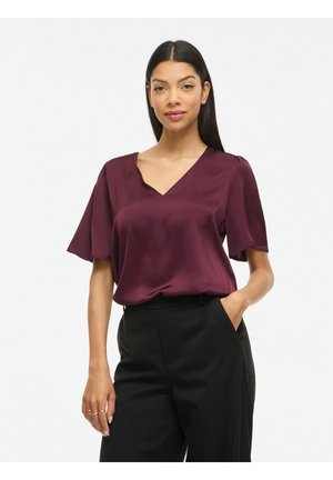 Femme aux longs cheveux foncés portant un chemisier bordeaux à manches courtes et un pantalon noir, debout avec une main dans la poche devant un fond blanc.