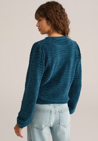 Teal-texturerad tröja med rund halsringning, puffärm och ribbad fåll. Buren med ljusblå jeans, vilket visar en avslappnad passform.