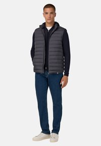 Gilet imbottito grigio con cappuccio, caratterizzato da un design trapuntato e chiusura con zip. Abbinato a una maglietta a maniche lunghe blu navy, jeans blu e sneakers bianche.