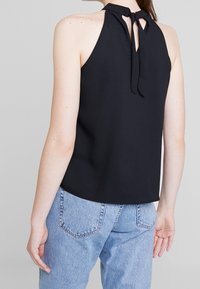 Haut noir sans manches avec un col montant et une fermeture à nouer dans le dos ; tissu lisse ; associé à un jean bleu clair taille haute.