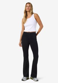 Calças pretas evasê com ajuste justo nos quadris, combinadas com um top branco canelado. O modelo calça ténis cinzentos. Fundo simples.