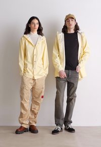 Două modele stau unul lângă celălalt. Unul poartă o jachetă de ploaie galbenă și pantaloni cargo bej; celălalt poartă un tricou negru și blugi gri.
