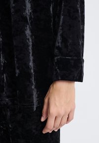Tela de terciopelo negro con una textura suave, que presenta un puño doblado y un corte relajado. La mano descansa sobre la tela, mostrando el detalle.