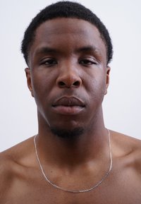 Eine silberne Kettenhalskette liegt auf einer nackten Brust und hebt ihre glänzende, glatte Textur gegen die glatte Haut hervor. Der Hintergrund ist rein weiß.