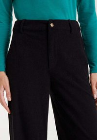 Pantalones de pana negros con una cintura alta, con un detalle de botón dorado. La tela tiene una textura acanalada y un diseño holgado de pierna recta.