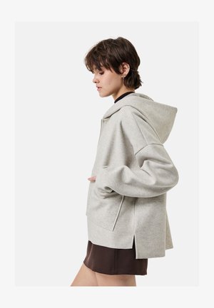 Nuori henkilö, jolla on lyhyet hiukset, pukeutuneena vaaleanharmaaseen hupulliseen takkiin ja tummaan hameeseen, poseeraa käsillään takin taskuissa.