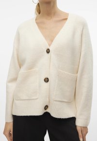 Krämfärgad cardigan i mjukt material, med djup V-ringning, tre stora knappar och två framfickor med ribbad struktur.