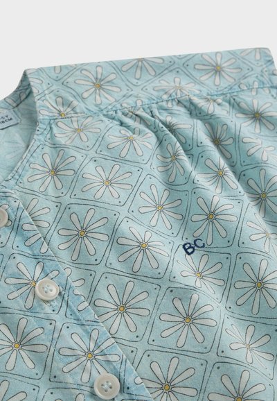 Chemise bleu clair avec motifs de marguerites blanches dans des contours carrés, centres jaunes, devant boutonné, et petit "BC" brodé sur le tissu.