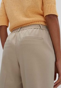 Beige elastische Bundhose mit zwei Gesäßtaschen, kombiniert mit einem kurzen, strukturierten orangen Strickoberteil. Sichtbare Nähte an der Taillenlinie.