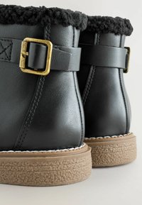 Schwarze Leder-Ankle-Boots mit einem strukturierten schwarzen Fellkragen, goldfarbenem Metallverschluss und einer tanfarbenen Gummisohle mit gerippten Details.