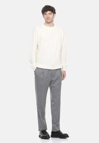 Uomo in maglione crema, pantaloni grigi a pieghe e mocassini neri, in piedi contro uno sfondo chiaro uniforme con le mani in tasca.